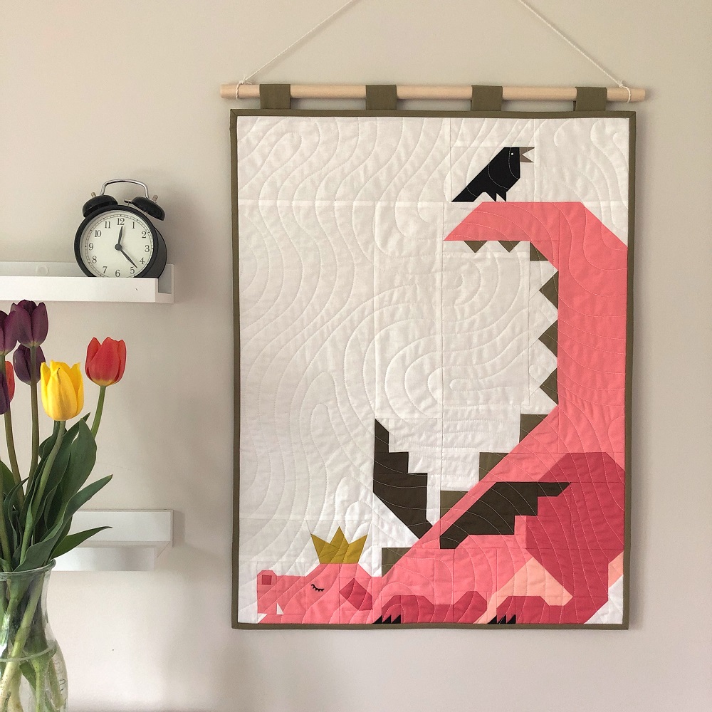 Quilted wall hanging featuring a pink dragon wearing a crown and a black bird sitting on its tail.