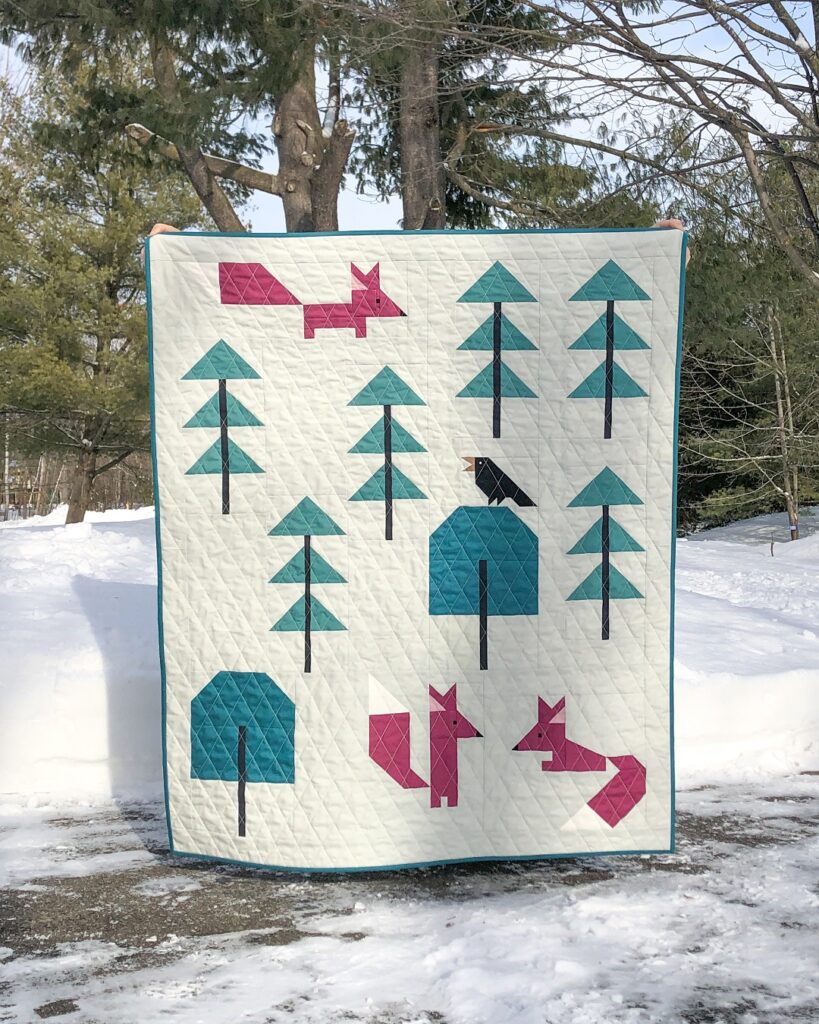 Apples & Beavers - Birds and foxes quilt tutorial: Crib quilt held up in front of snowy evergreen trees